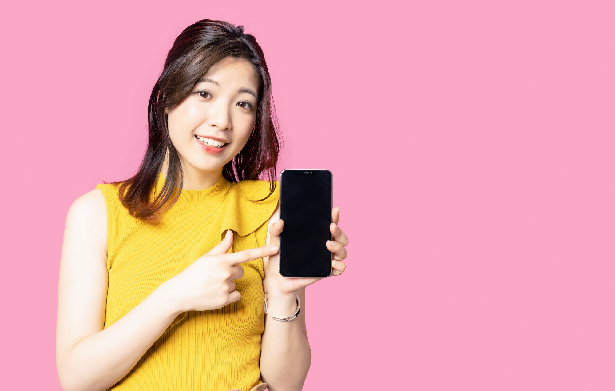 ピンク色の壁を背景にスマートフォンを指さしている女性